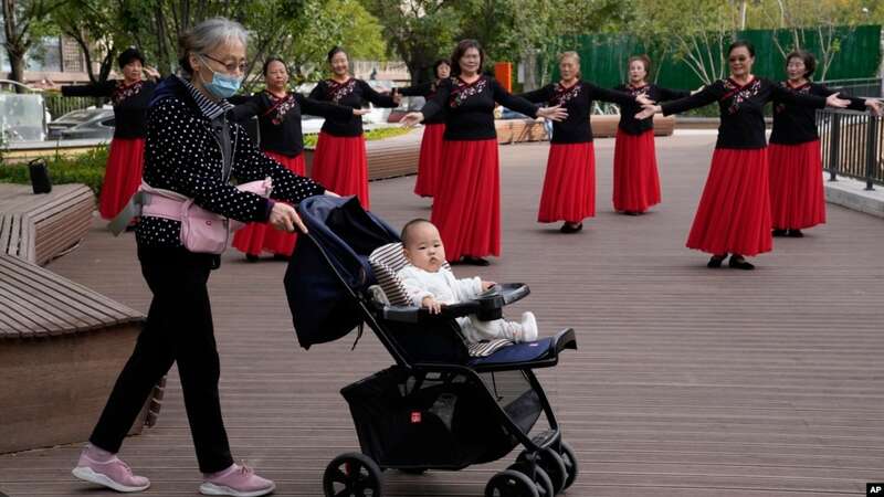 一名老年妇女推着一个婴儿走过北京一所公园里跳广场舞的妇女